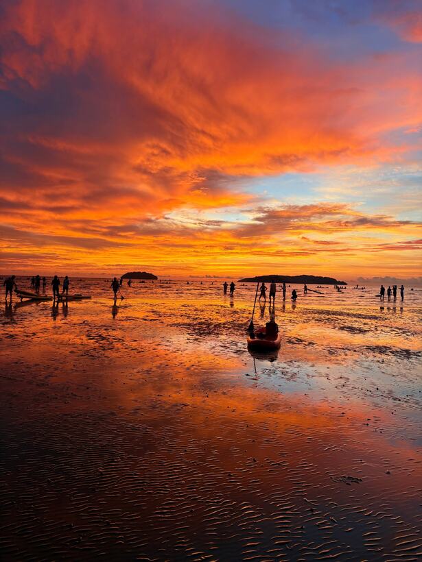 Low tide at sunset: an orange and red sky mirrored in the wet sand, a kayak and paddlers in silhouette