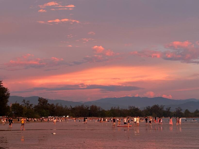 A coral and pink sky over a wide tidal flat with hundreds of people standing in the shallow water, mountains behind