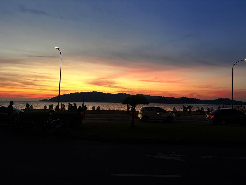 An orange and blue sunset over a bay with two islands, people and parked cars along the waterfront road
