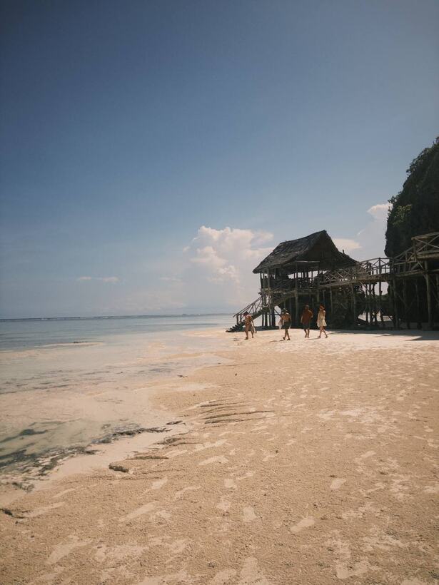 A wide white sand beach with very shallow clear water and a thatched hut on stilts
