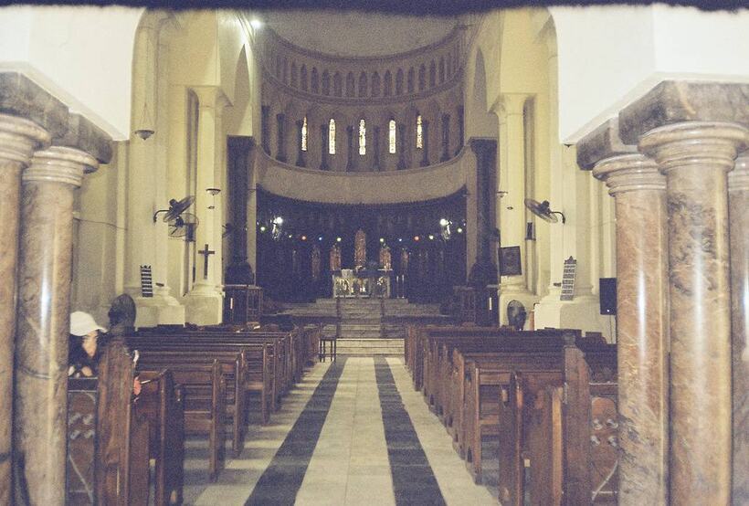 The nave of a church: marble columns, wooden pews and a striped stone floor leading to a lit altar